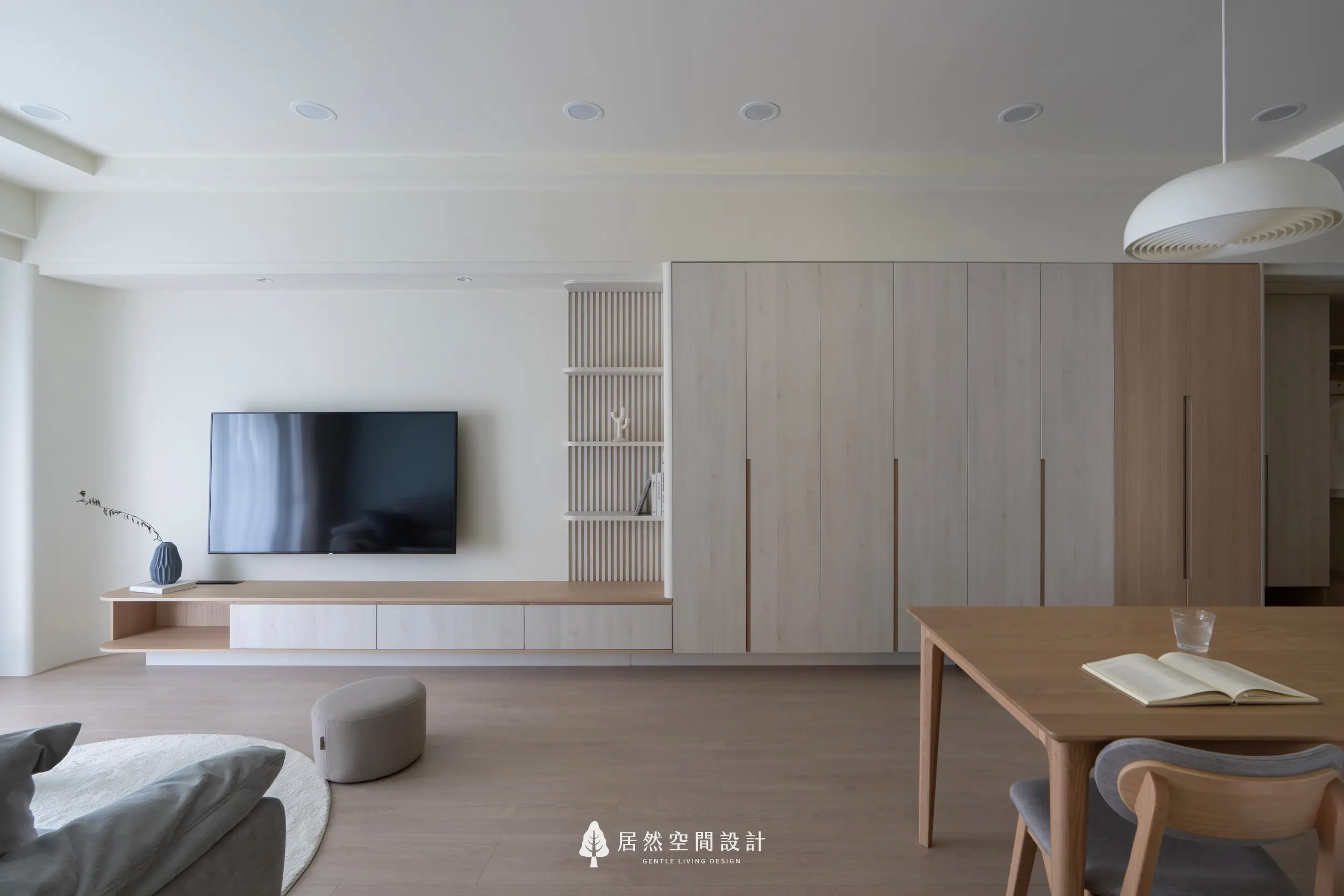 開放式客餐廳以輕柔白色與淺木質串聯整體視覺，電視主牆結合層板與大量收納，；前景木質餐桌延伸生活場景，使空間展現舒適流動的居家節奏。