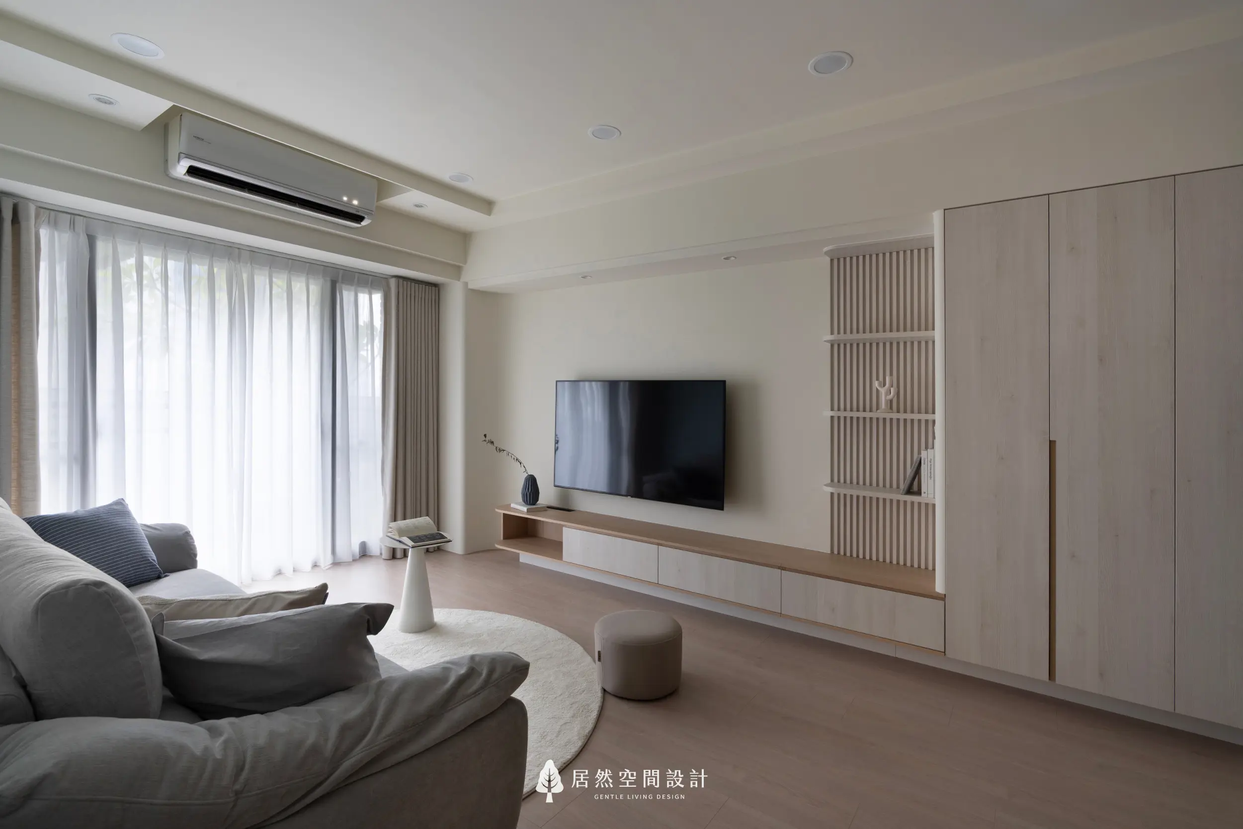 客廳以柔白與淺木色鋪陳開闊視覺，電視主牆結合層板與收納櫃；落地窗引入自然光，搭配舒適沙發與圓形地毯，營造放鬆且暖意柔和的生活場景。