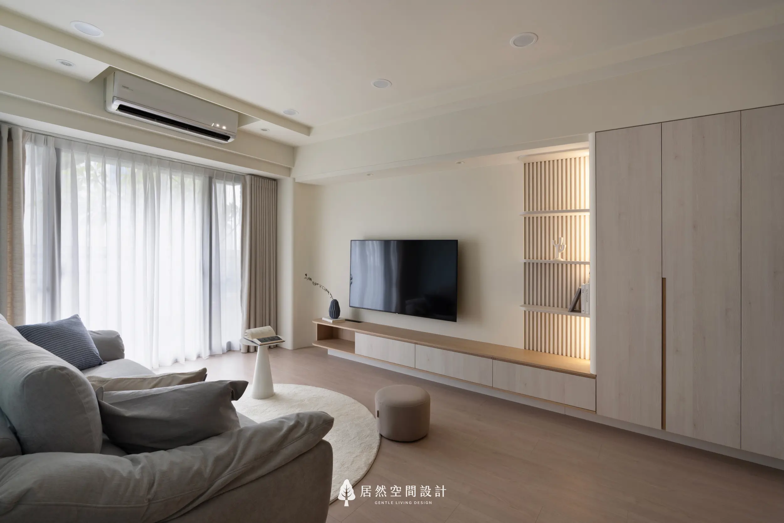 客廳以大片窗簾引入柔光，米白電視主牆搭配木質收納層板，營造清爽溫潤的氛圍；舒適沙發與圓形地毯構成放鬆角落，呈現恬靜而自在的日常感受。