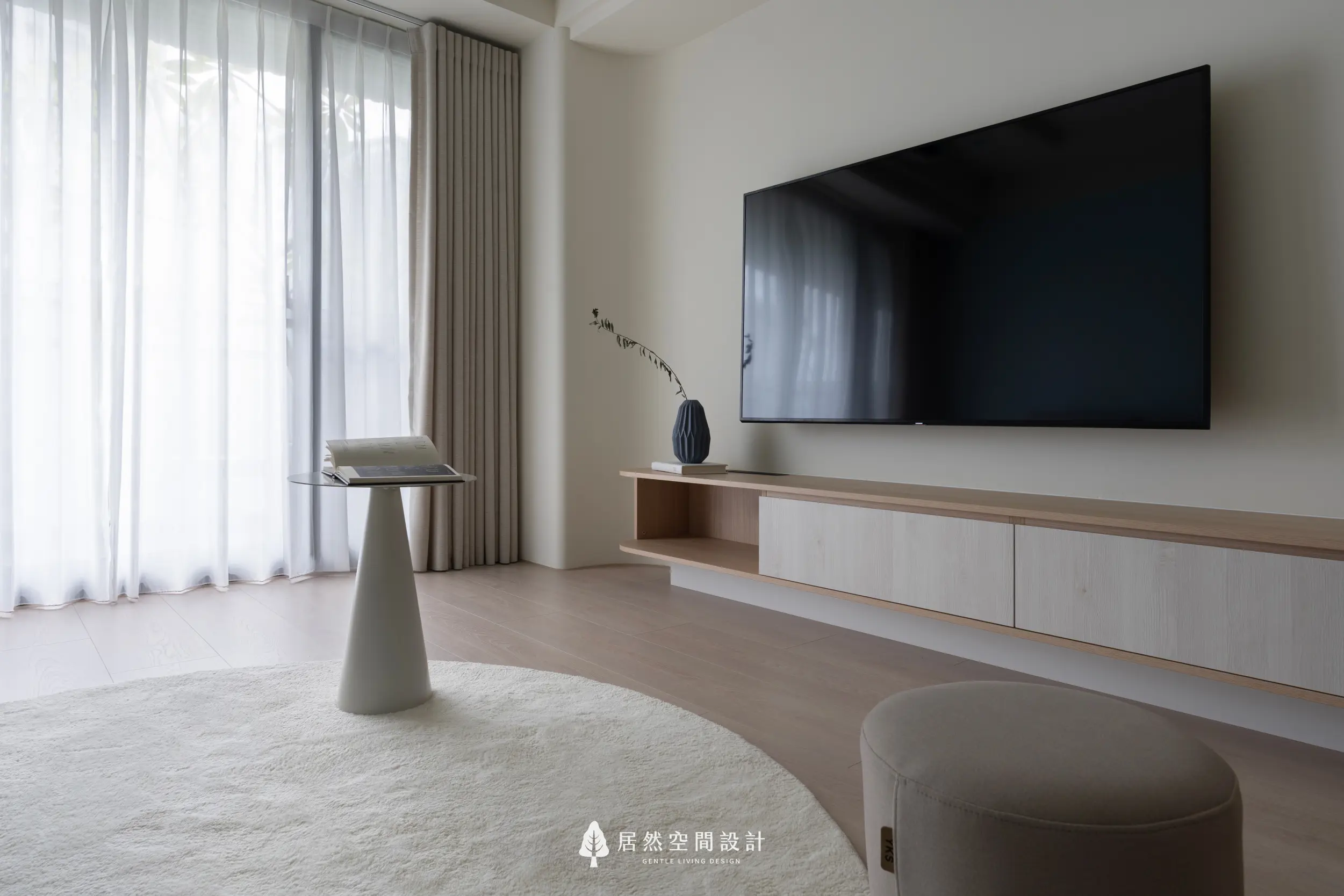 落地窗透入柔和自然光，照亮客廳的米白地毯與極簡邊几；電視主牆以淺木色櫃體延伸輕盈視覺，營造安靜放鬆的舒適日常場景。