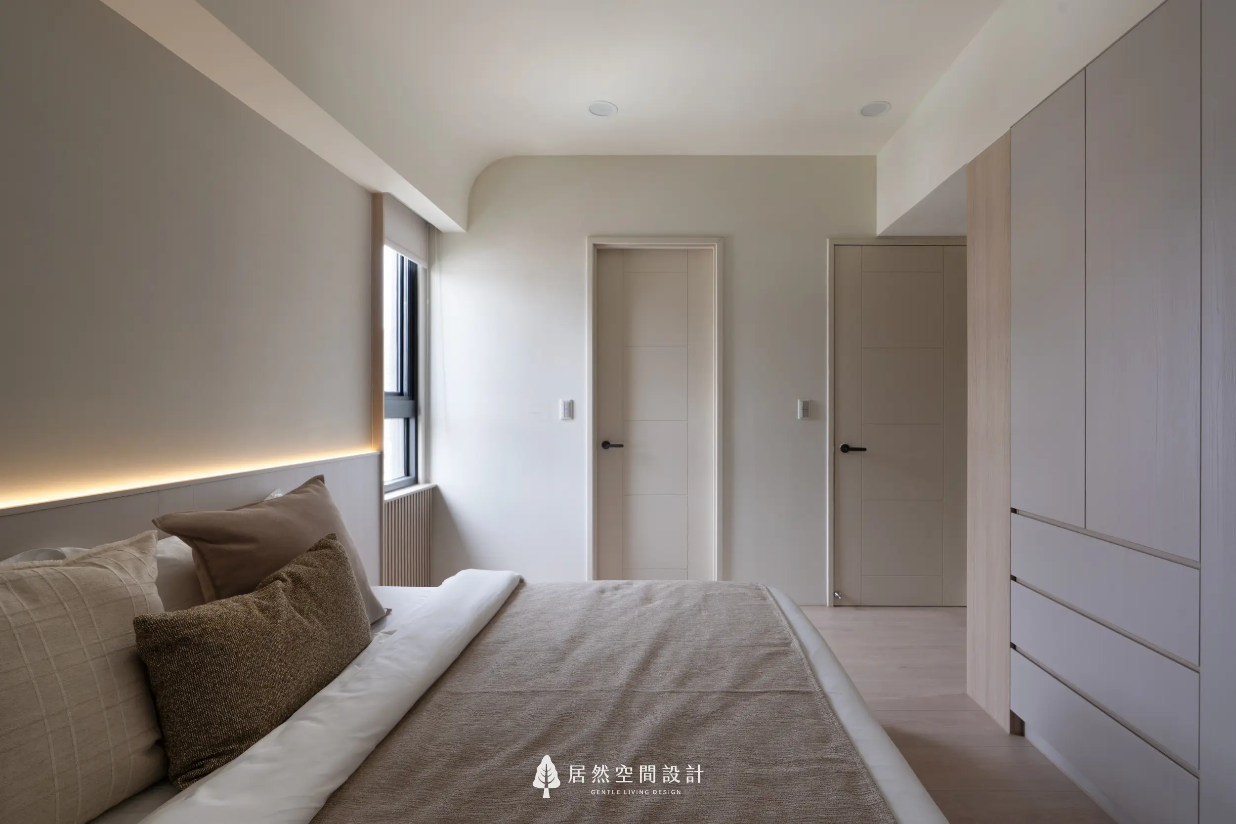 主臥床頭加入柔和間接照明，讓淺米色與木質調更顯溫暖；自然採光自側窗引入，右側收納櫃體維持整潔，營造安定舒適的休息氛圍。