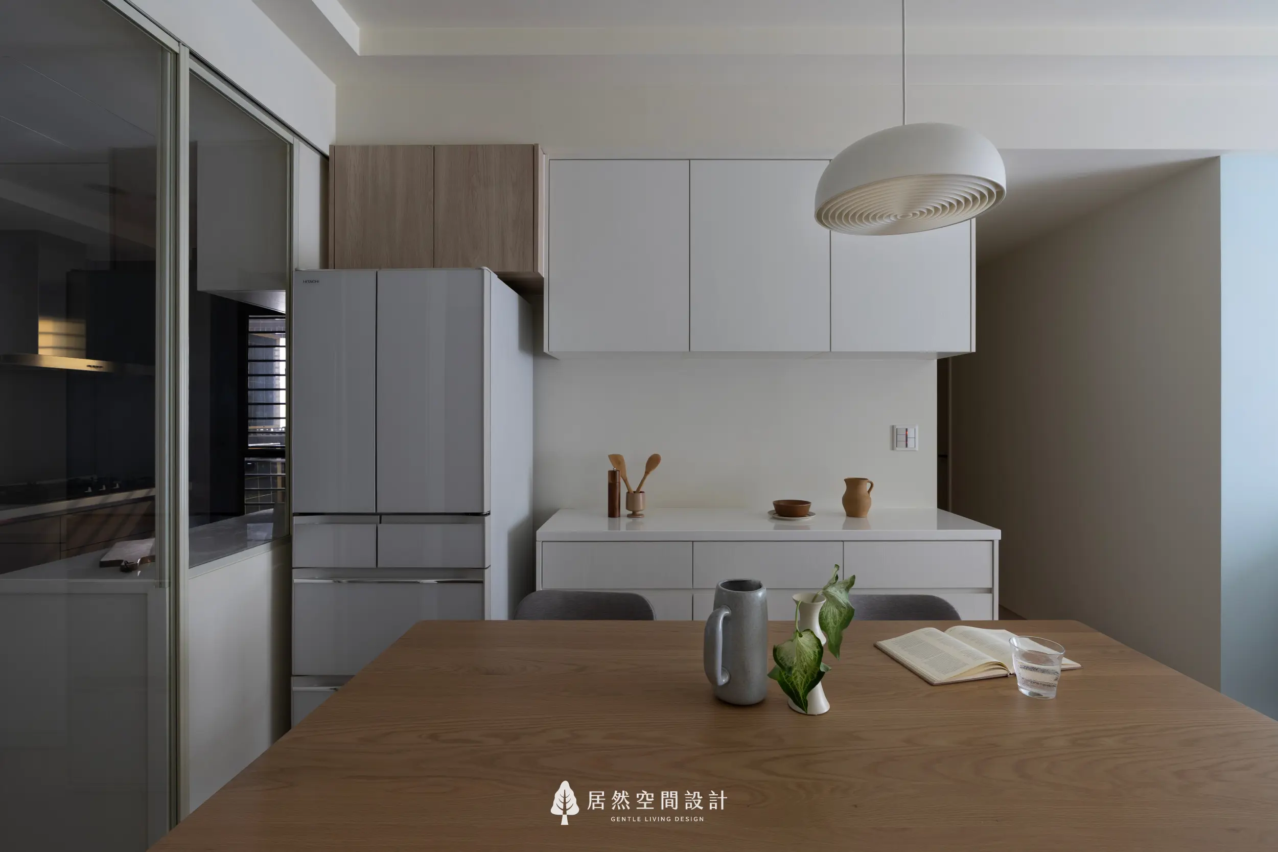 白色上櫃與淺木色元素交織出清爽廚房，冰箱與備餐檯配置緊湊但井然有序；前景餐桌擺放書本與花器，為日常注入悠緩節奏。