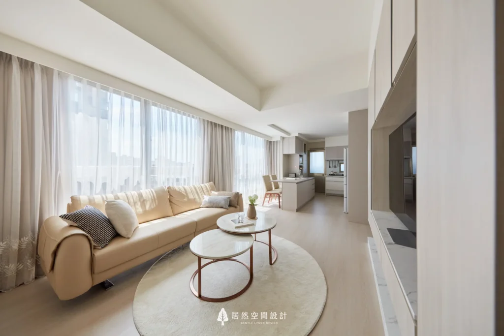 客廳沿窗配置米色沙發，搭配圓形茶几與地毯，柔化動線尺度，長向格局串聯餐廚空間，透過自然採光營造明亮舒適的日常生活氛圍