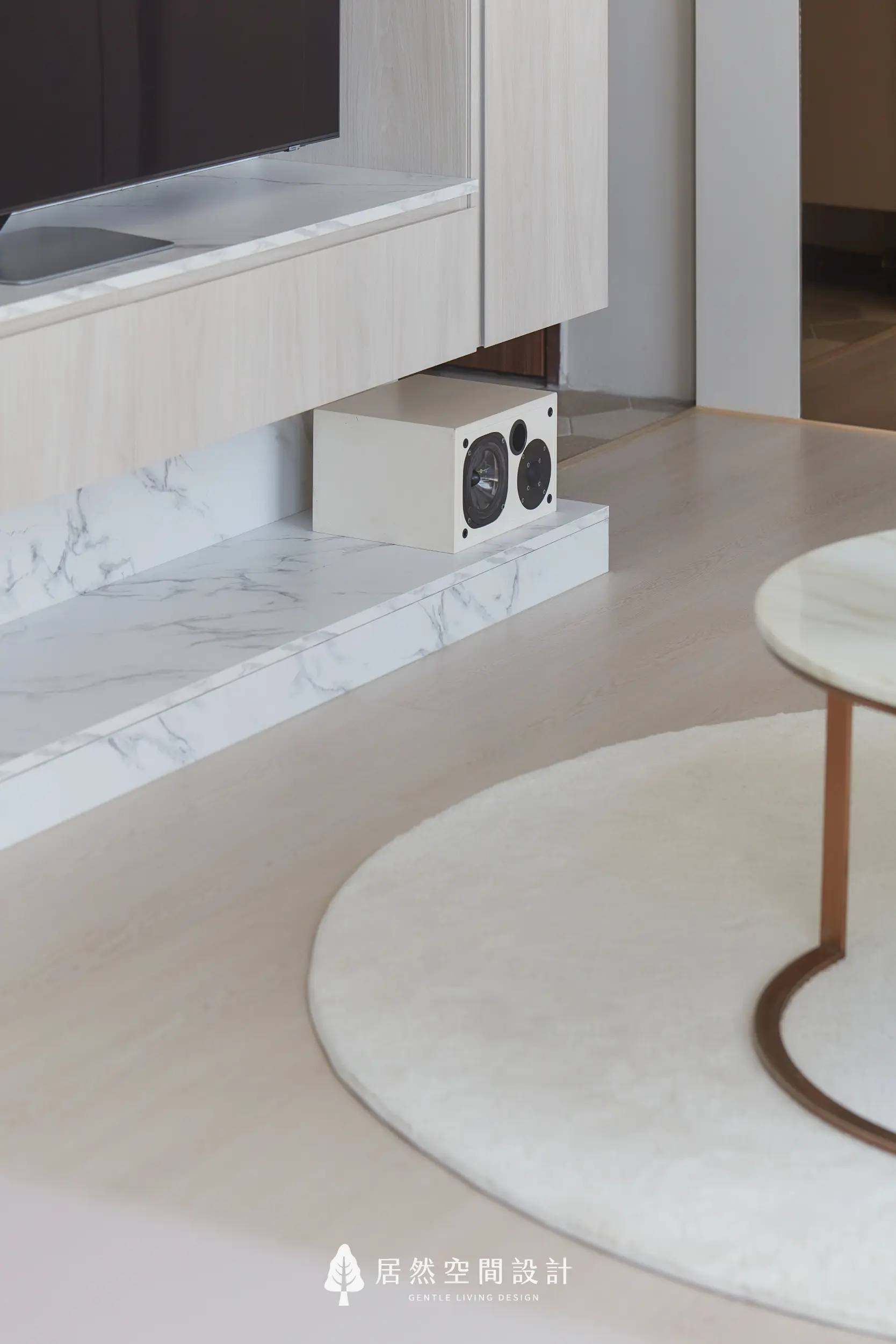 電視牆下方以石紋平台整合音響設備，線條俐落收整視覺，搭配淺木櫃體與圓形地毯，維持客廳機能完整與整體畫面清爽平衡