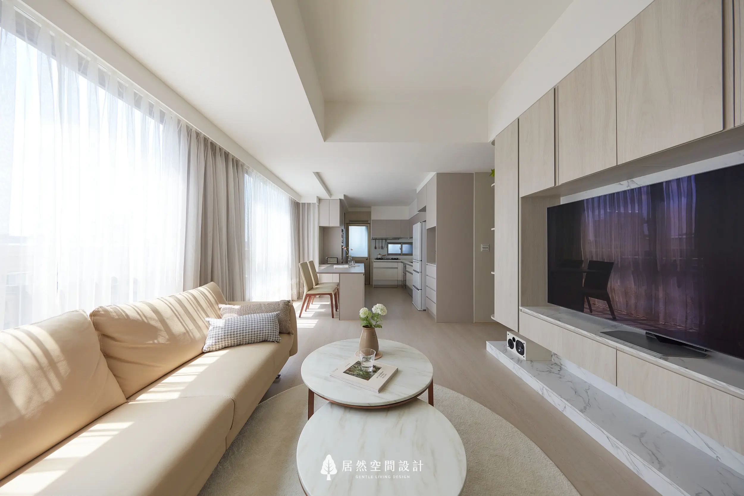 客廳採長向格局串聯餐廚空間，電視牆結合淺木櫃體與石紋平台，搭配米色沙發與圓形茶几，引入自然光線，形塑開闊流動且溫潤的生活尺度
