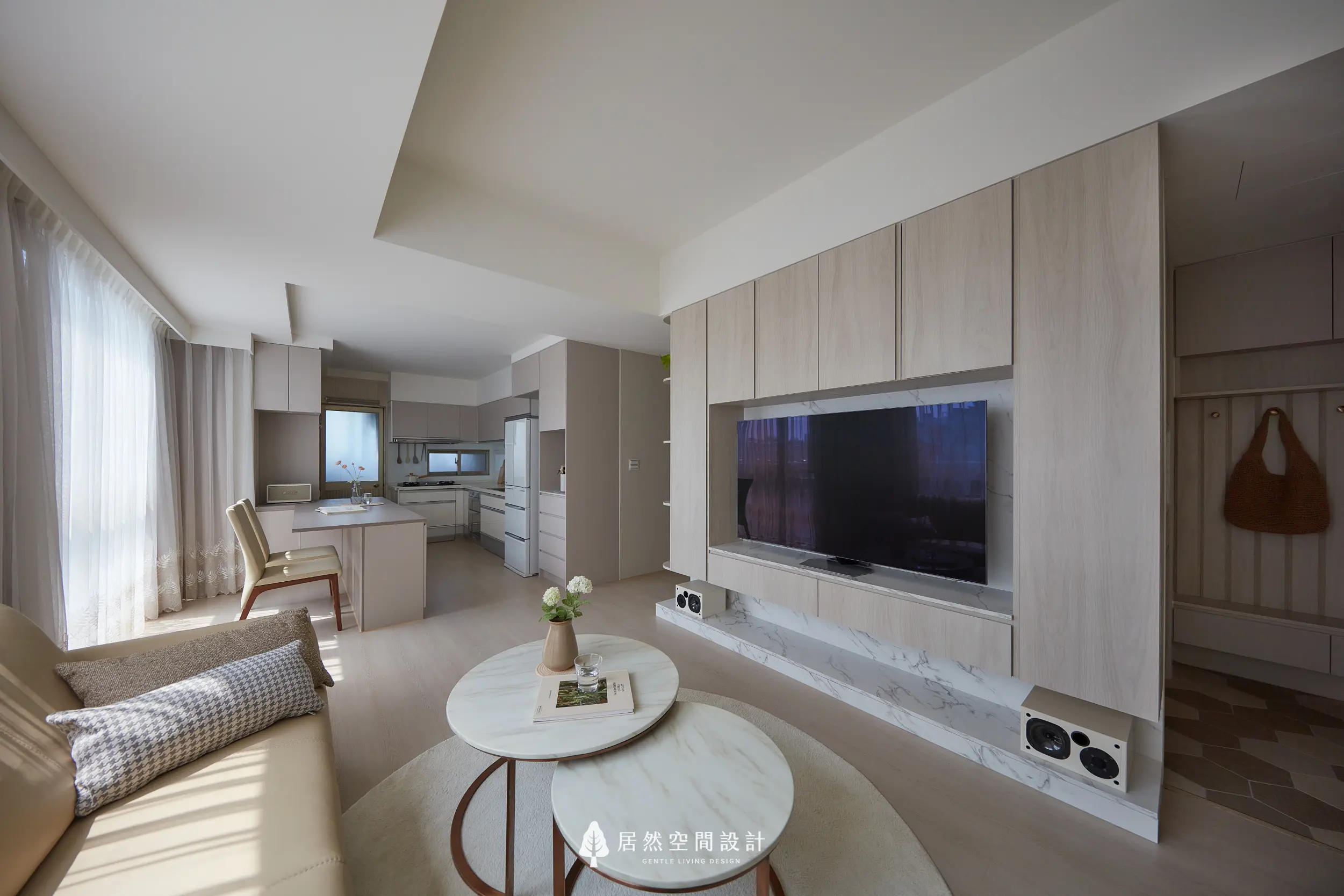客廳以淺木電視牆結合石紋檯面，串聯餐廚空間，圓几與柔光配置，形塑通透開放的日常生活場景，延伸採光與收納層次，營造溫潤平衡居家氛圍