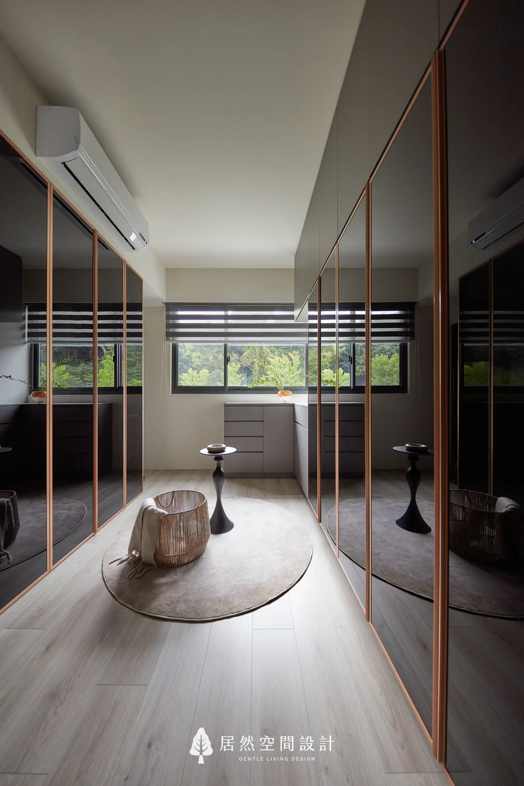 更衣室以雙側玻璃衣櫃與金屬框架打造通道式收納，中央圓毯與小邊桌形成展示區，窗邊自然採光與綠景映入室內，營造沉靜開闊的更衣空間