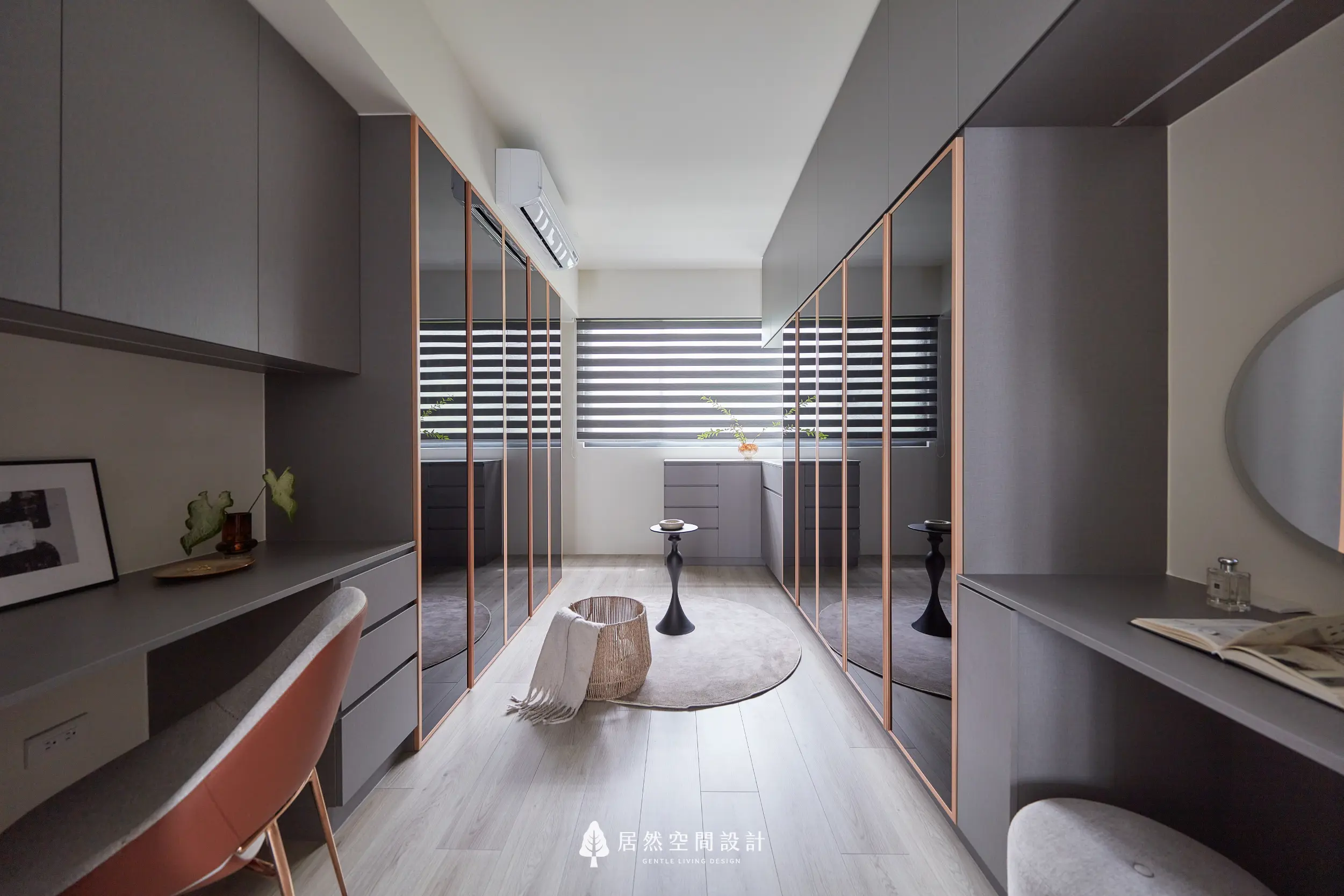 更衣室採雙側玻璃衣櫃打造通道式收納動線，結合吊掛區與抽屜櫃機能，中央圓毯與小邊桌形塑展示氛圍，窗邊自然採光提升整體空間感