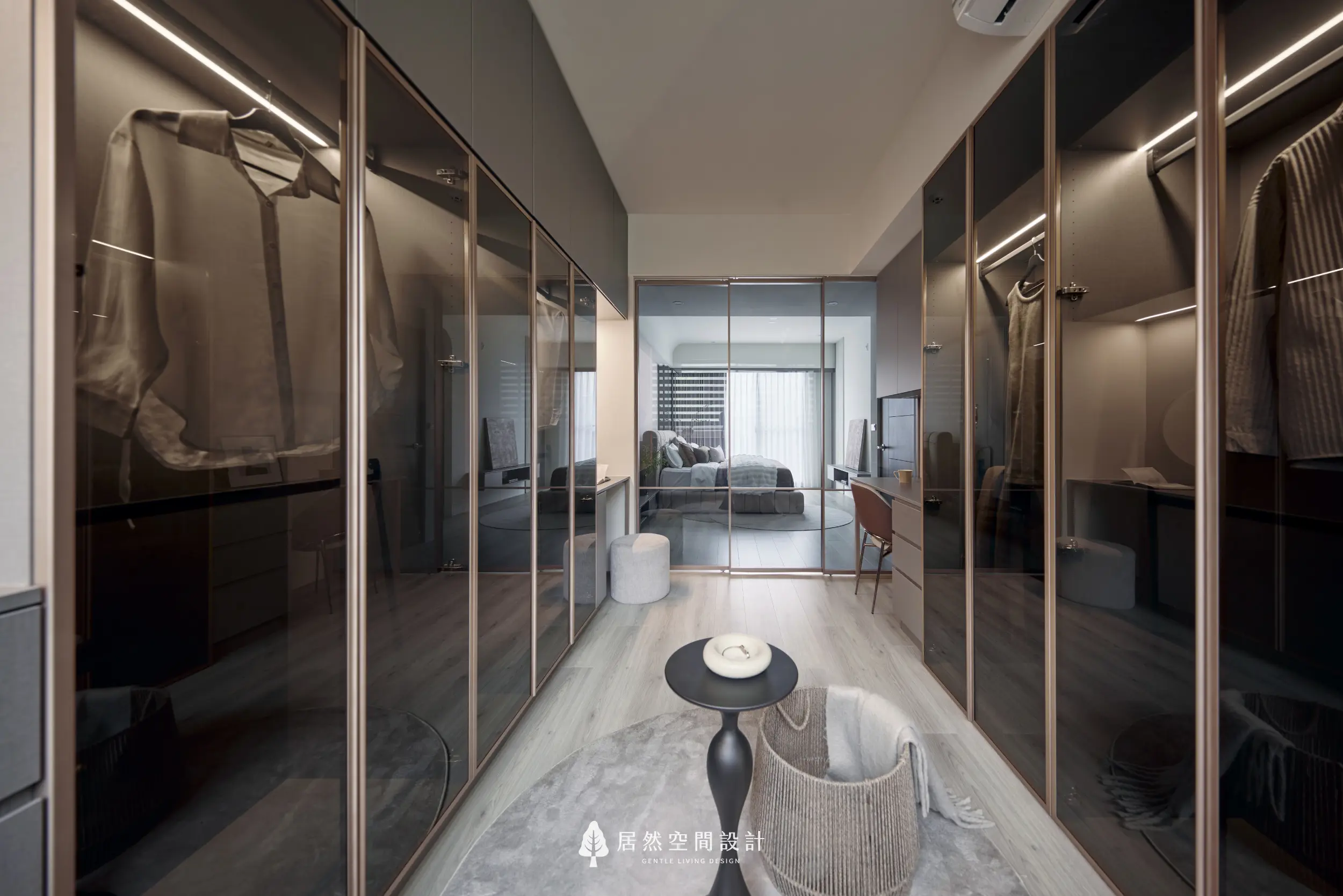 更衣室以雙側玻璃衣櫃形成通道式動線，結合吊掛收納與間接燈光展示衣物，視線延伸至主臥室窗景，營造通透寬敞的更衣空間層次感
